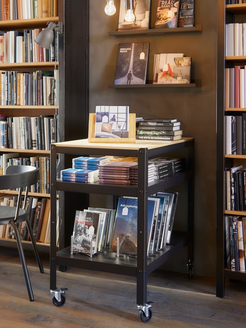 A beige trolley with a tablet stand and containing multiple books is placed between two bookcases, with picture ledges above.