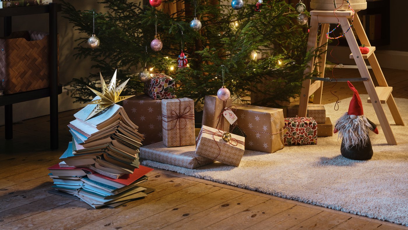 Julgran med ljusslingor och julgranskulor i ett mysigt vardagsrum, omgiven av inslagna julklappar och böcker.