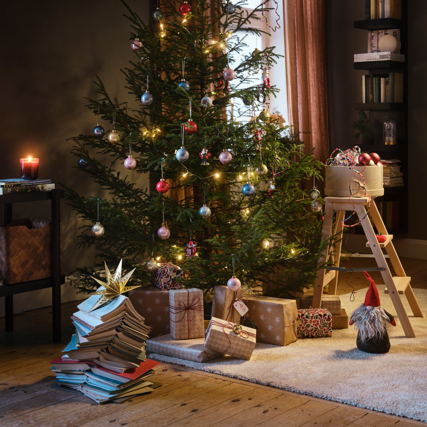 Julgran med ljusslingor och julgranskulor i ett mysigt vardagsrum, omgiven av inslagna julklappar och böcker.