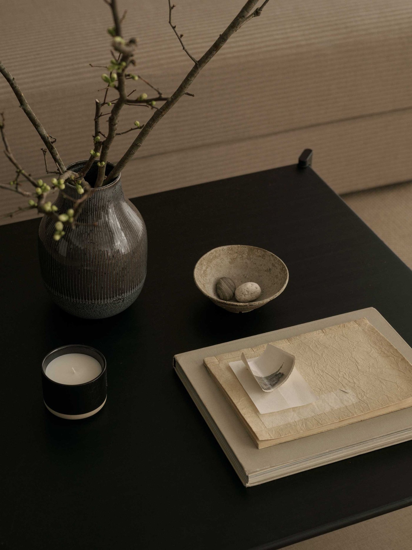 A beautifully styled dark surface, perfect for adding a touch of natural elegance and calm to your home. A ribbed, dark grey vase holds delicate branches with fresh green buds, bringing a hint of the outdoors in. Nearby, a small, textured bowl cradles two smooth pebbles, adding a touch of natural serenity. A cosy candle in a black holder with a white base promises a warm glow for a relaxing evening. A stack of books and papers, topped with a small, curved decorative object, suggests a quiet moment for reflection. This arrangement creates an inviting and peaceful atmosphere, showcasing how thoughtful details can transform a space.