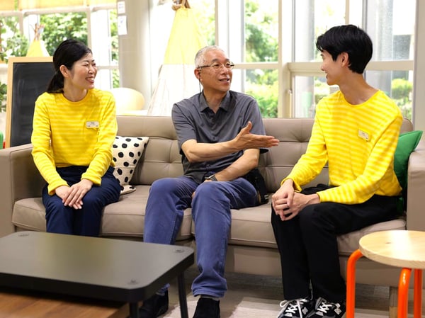 黄色のストライプシャツの2人と中央の人物が、イケアのベージュのソファに座って会話をしている。