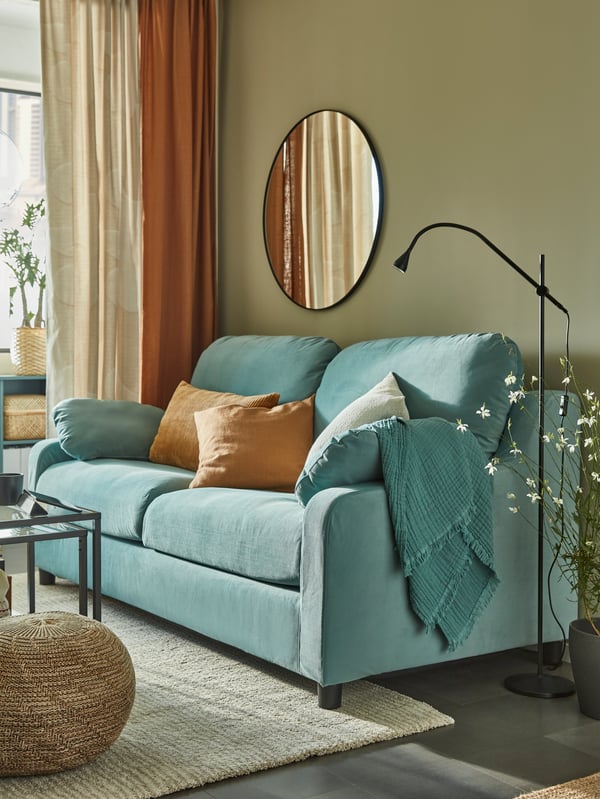 A light turquoise high-back sofa against a moss green wall, a round mirror, dark beige cushions, a black reading lamp.