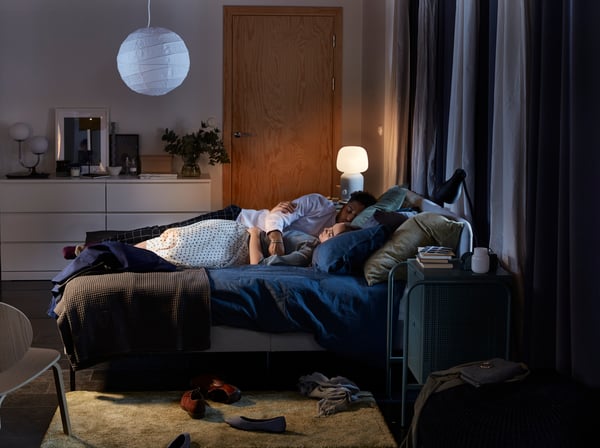 A couple cuddle asleep in a SLATTUM upholstered bed, with a SYMFONISK lamp/speaker lit in the background.