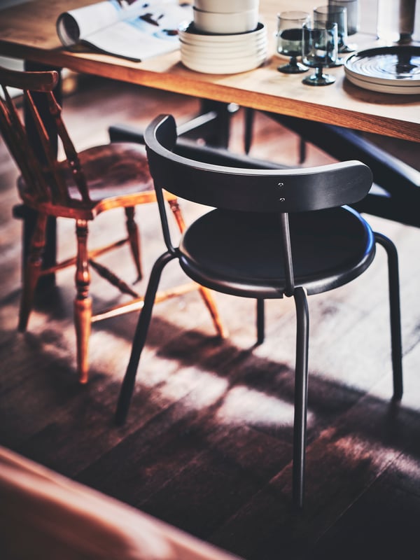 A dining area with a black YNGVAR chair and a brown, wooden chair set at a SKOGSTA dining table with tableware items.
