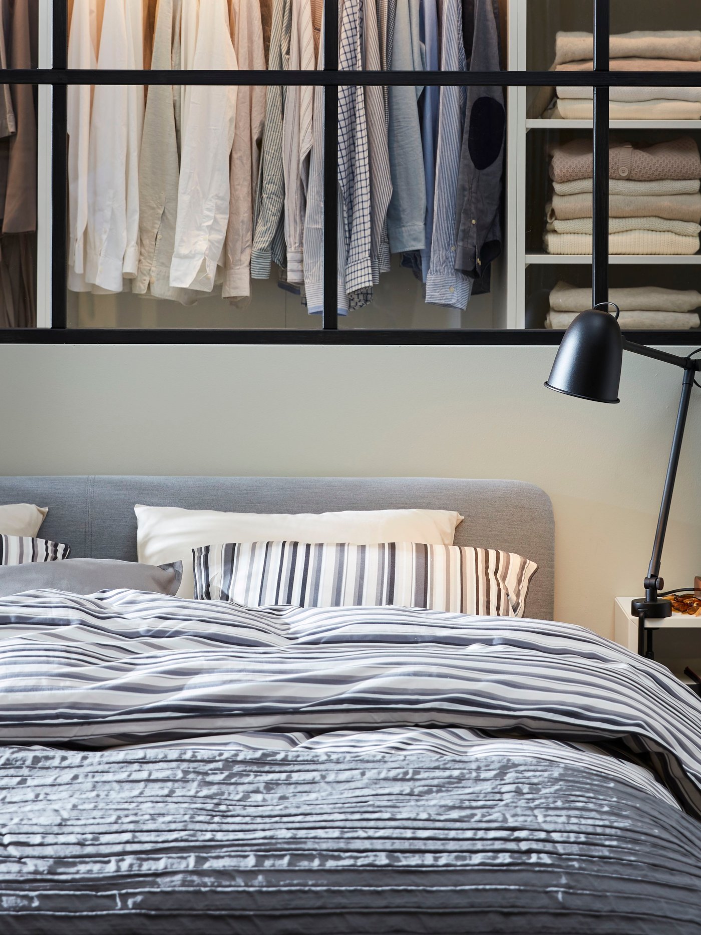 Szara, tapicerowana rama łóżka, tekstylia w paski, biały stoik nocny i czarna lampa.