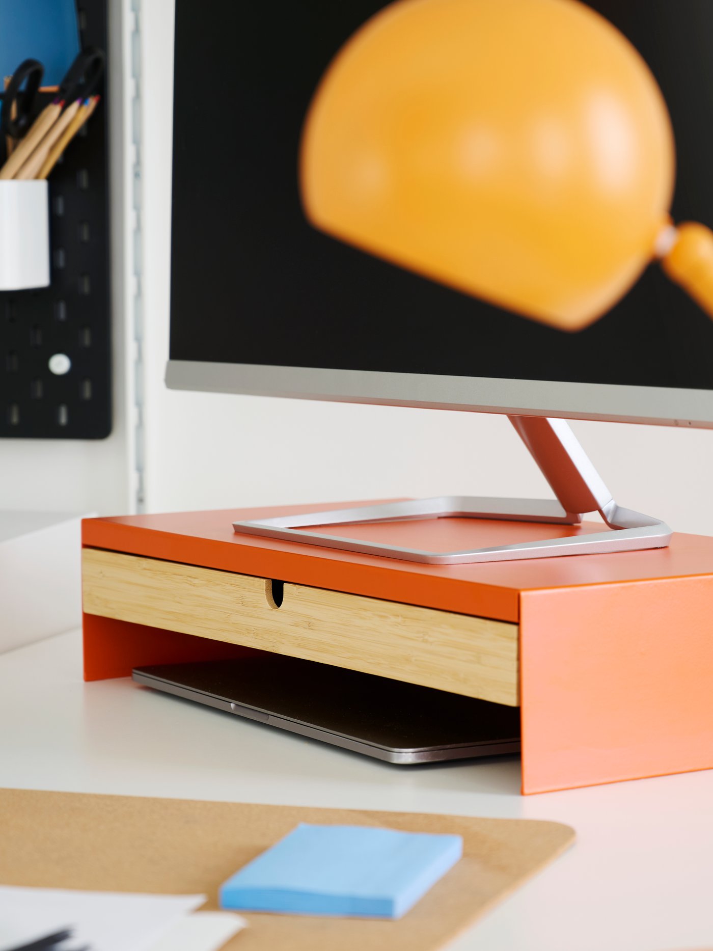 A close-up view of a bright orange ELLOVEN monitor stand with a drawer, placed on a desk next to a computer.