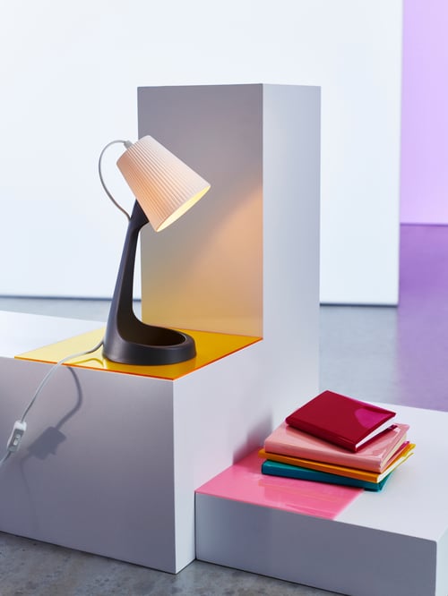 A SVALLET work lamp in dark grey and white on a display stand, overlooking some diversely-coloured books stacked in a pile.