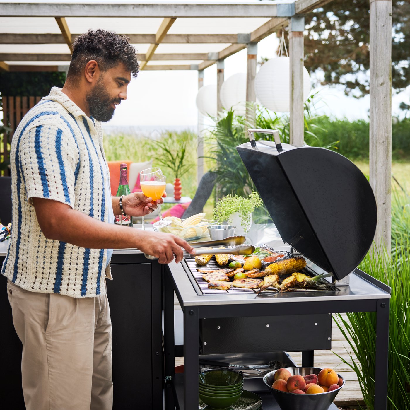 A patio with lush greenery features a black GRILLSKÄR charcoal barbecue made of black and stainless steel cooking food.