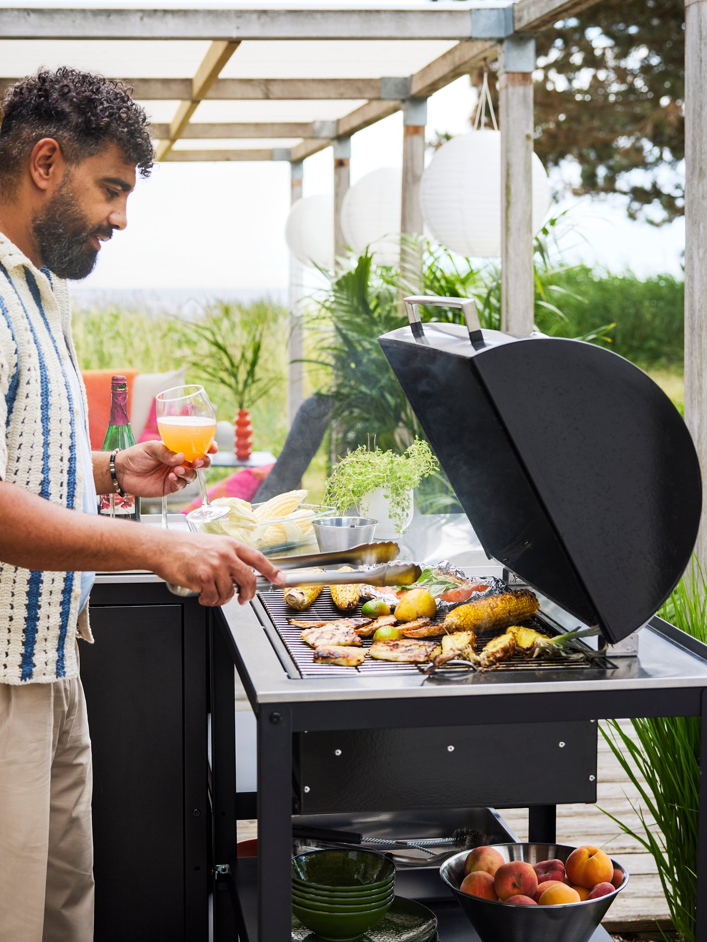 A patio with lush greenery features a black GRILLSKÄR charcoal barbecue made of black and stainless steel cooking food.