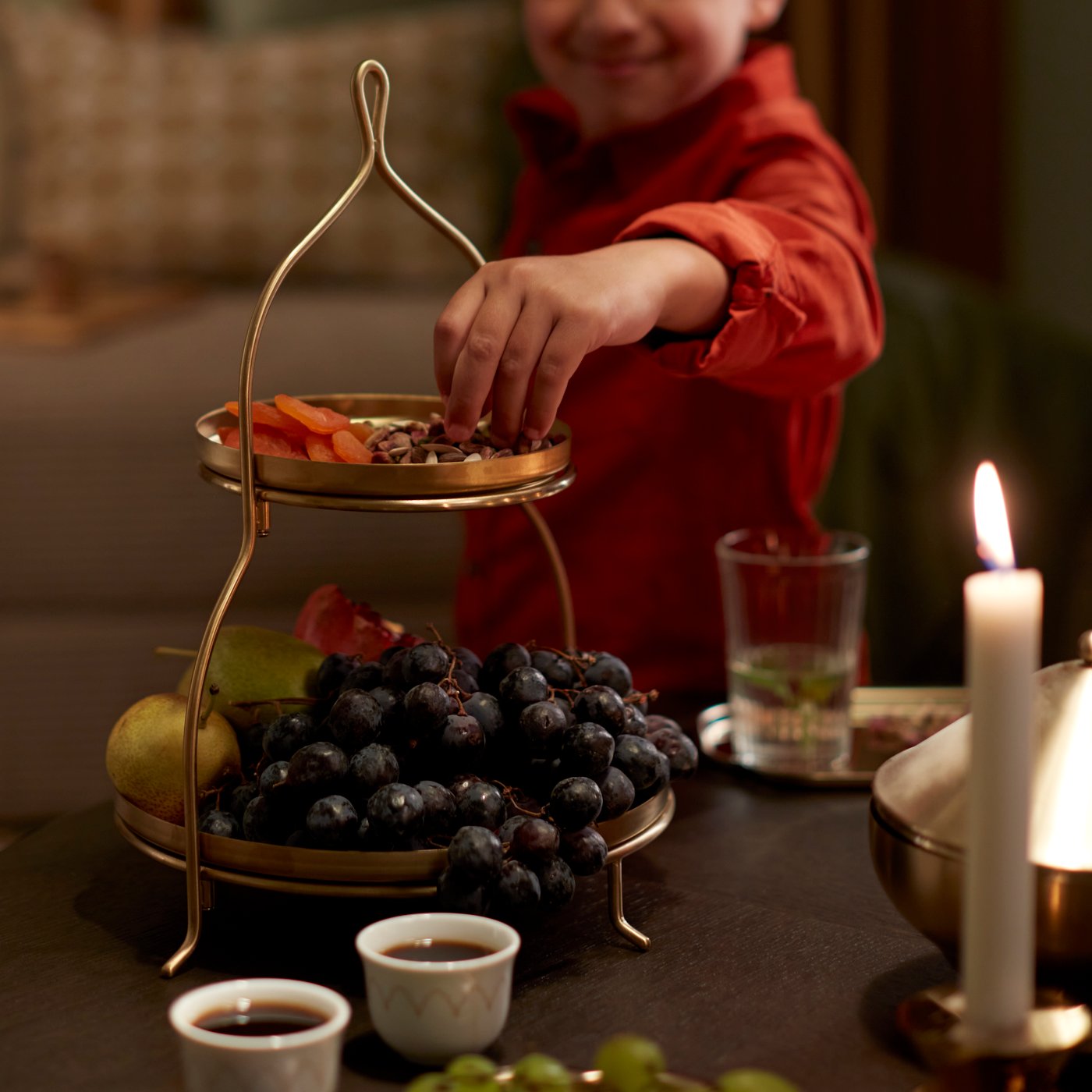 A child is reaching for some dates from a gold-coloured GOKVÄLLÅ two-tier serving stand with fruit and snacks.