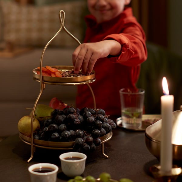A child is reaching for some dates from a gold-coloured GOKVÄLLÅ two-tier serving stand with fruit and snacks.