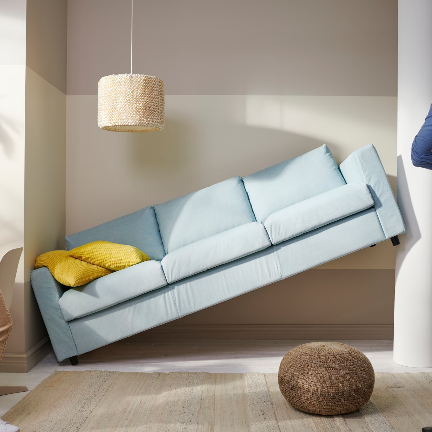 Para stoi naprzeciwko błękitnej sofy, która jest przechylona pod kątem, opierając się o ścianę i kolumnę w nowoczesnym, jasnym wnętrzu z żółtą poduszką na sofie i plecionym pufem na podłodze.
