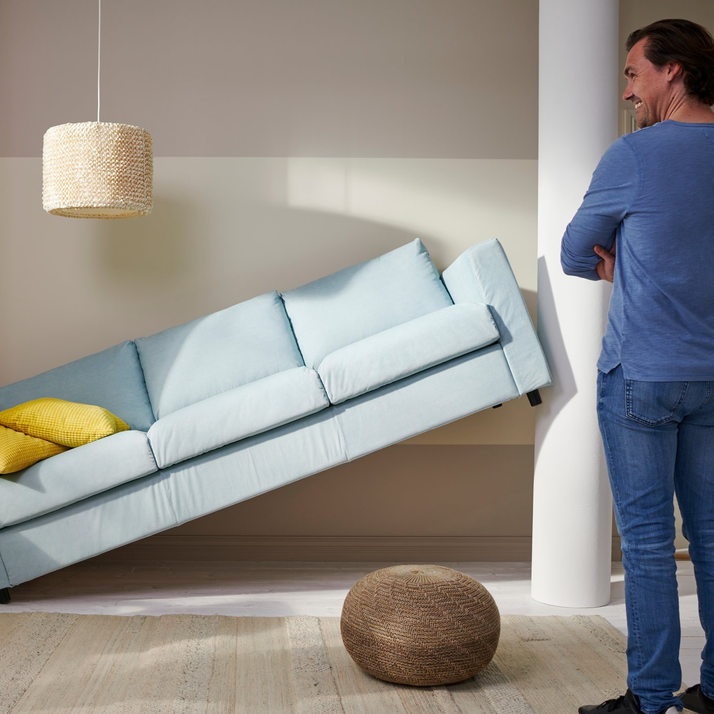 A smiling man with arms crossed, looking at a light blue sofa with yellow cushions that doesn't fit in their space.