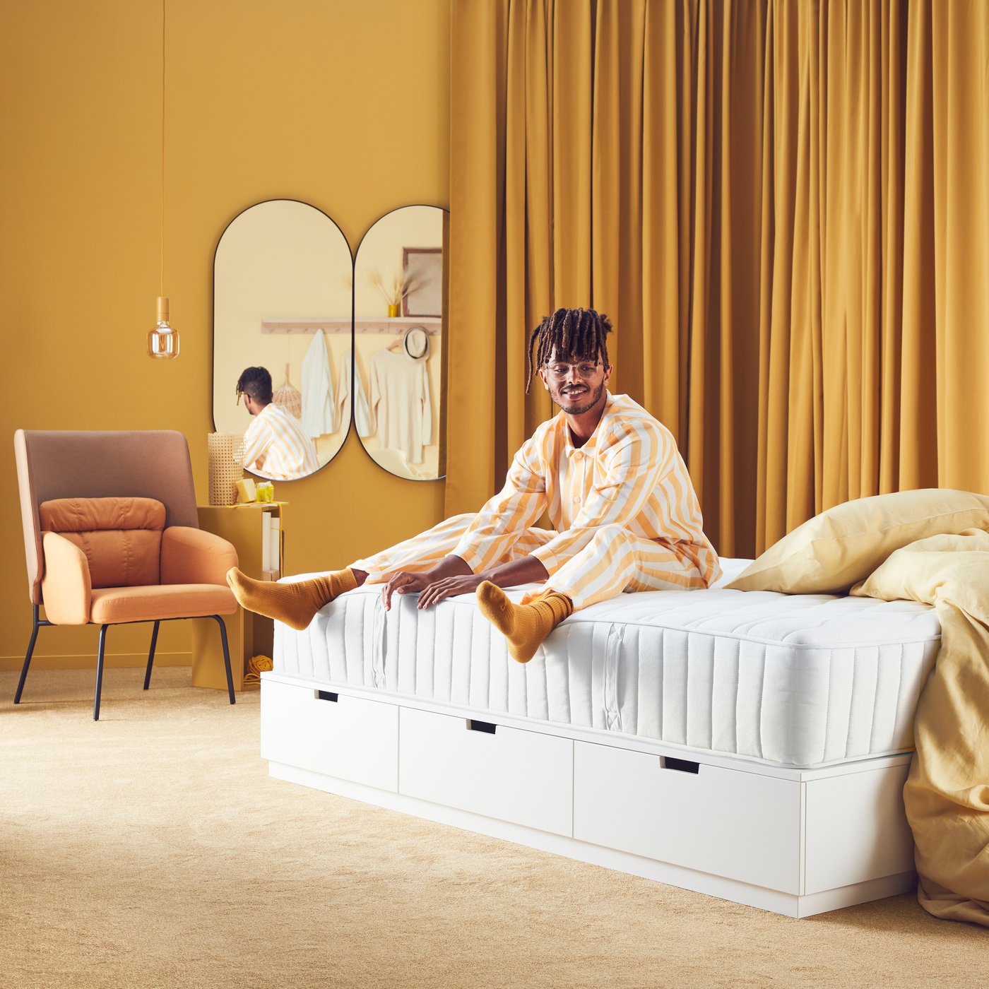 Un homme est assis sur un matelas blanc sur un lit dans une chambre aux rideaux brun doré SANELA pièce assombrissants.