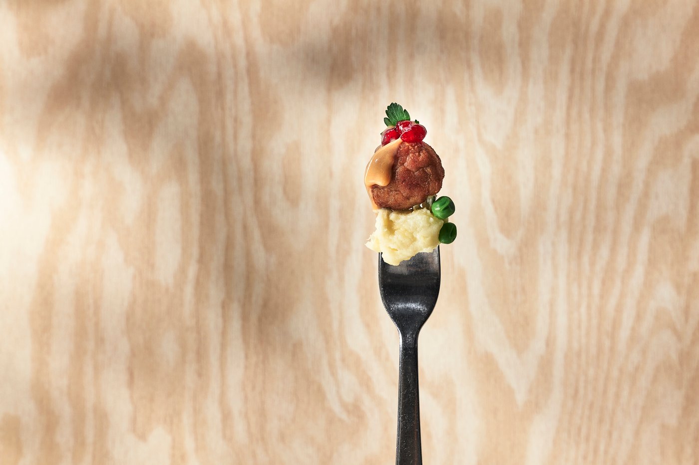 A fork with mashed potatoes, peas, and a meatball topped with lingonberry sauce and gravy against a birch background. 