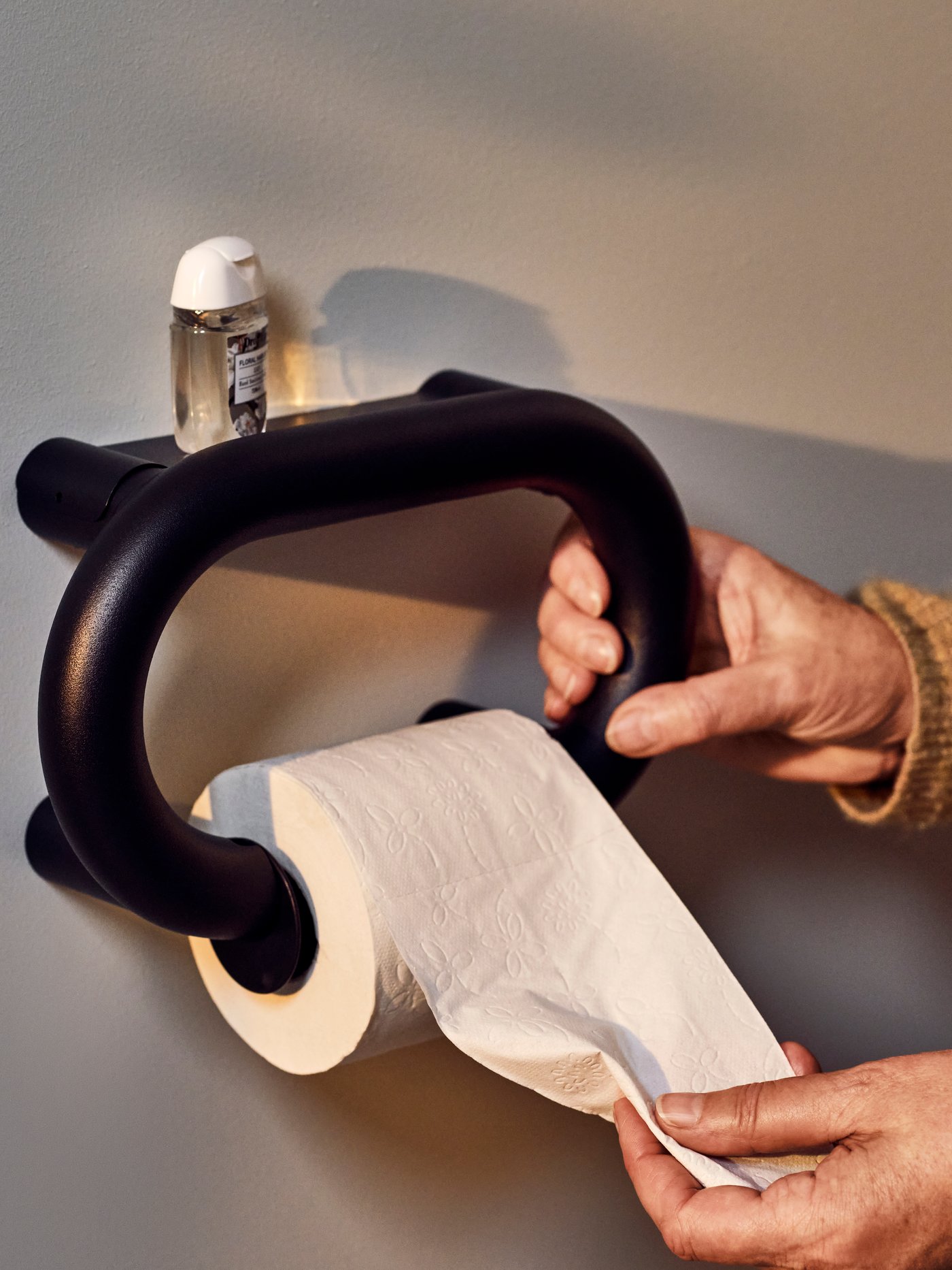 A person is holding onto a BÄSINGEN toilet roll holder in anthracite with one hand and taking some paper with the other hand.