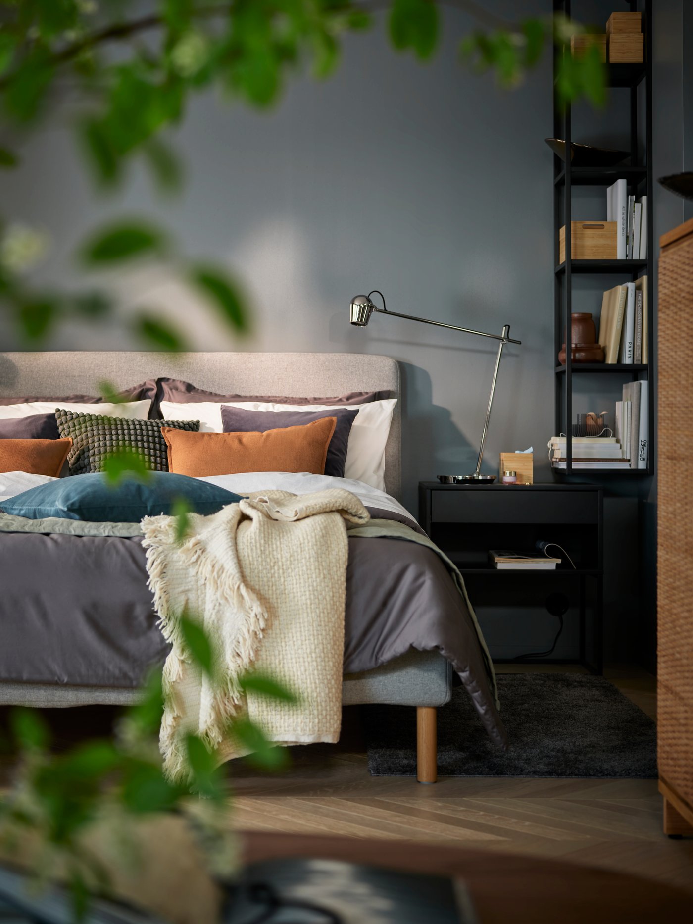 A bedroom with grey walls and view of bed, bedside table and shelves. Blurry green leaves from a plant in the foreground.