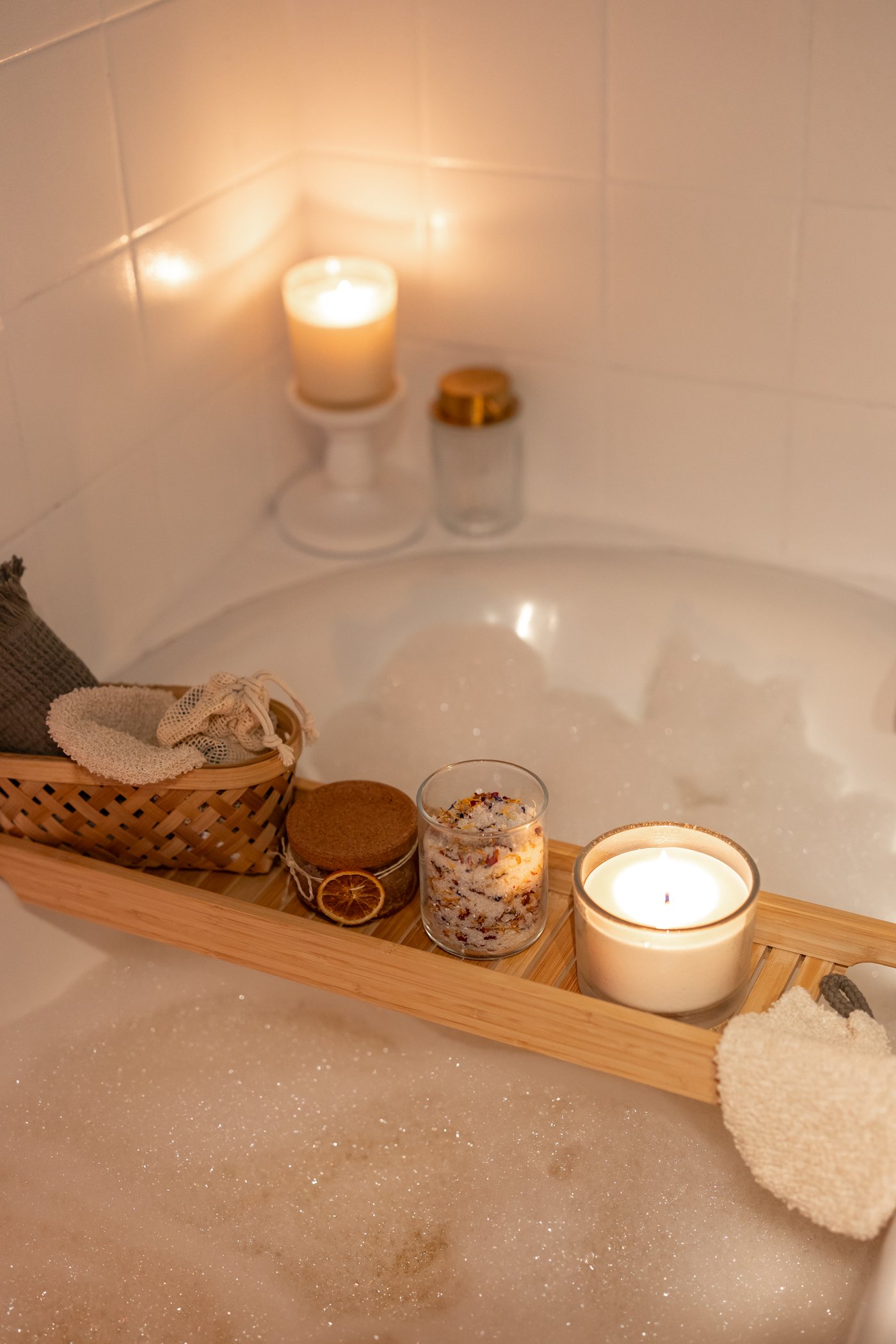 A bamboo HAVERN bath rack holding a lit scented candle, cotton pads, and potpourri above a bathtub filled with water and bubble bath.