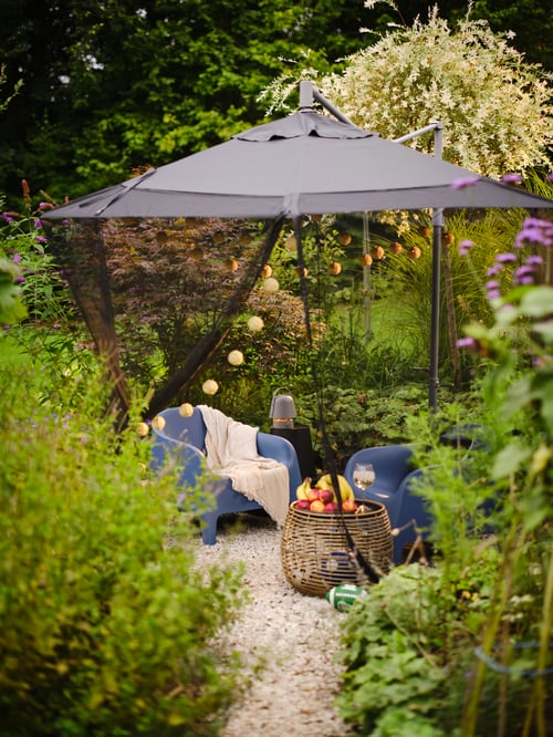 An outdoor area with a TORPÖN net placed on a hanging parasol with outdoor armchairs underneath and an outdoor table.