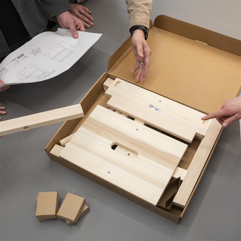A close-up of hands arranging parts from a wooden furniture assembly kit alongside instruction sheets on a grey floor.