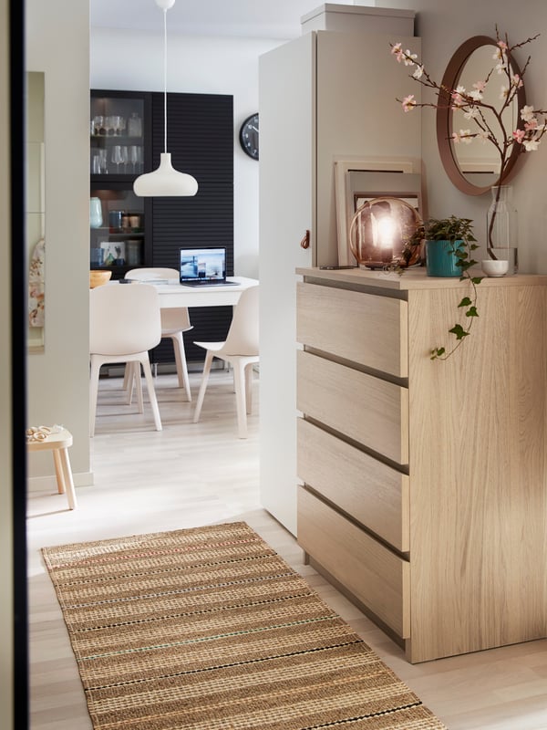 En MALM träbyrå med spegel, växt och dekorationer står i en korridor med vävd matta. I bakgrunden syns en matplats med stolar, ett bord med en laptop ovanpå, samt mörka skåp och hyllor längs väggen.