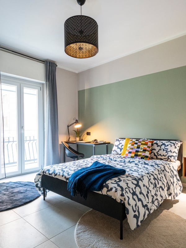 Una camera da letto con una struttura letto con federe e copripiumino a trame geometriche e con fantasie variegate. Sotto il letti due tappeti STOENSE e SKRIFTSPRÅK che attutiscono i rumori. A sinistra del letto, un angolo studio mentre alla destra un mobile ENHET a parete.