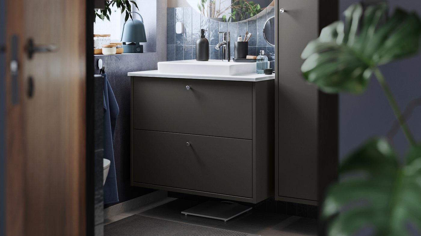 Un bany petit amb un armari de lavabo HAVBÄCK amb dos calaixos en gris fosc, un lavabo ORRSJÖN blanc i un mirall FAXÄLVEN rodó.