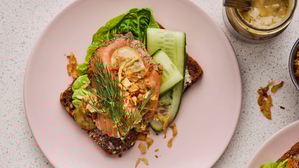A pink plate has a dark piece of rustic bread with several slices of salmon, lettuce, cucumber, dill and a mustard sauce.