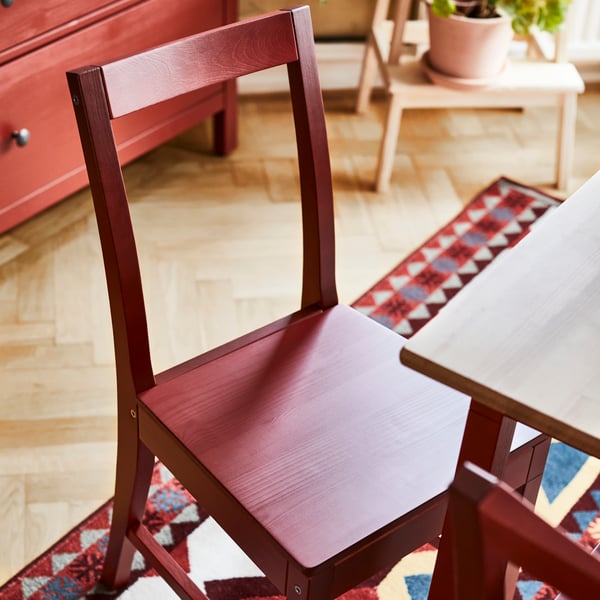 Roter Holzstuhl neben einem Holztisch; der Stuhl weist einen flachen Sitz und eine gerade Rückenlehne auf; auf dem Holzboden liegt ein gemusterter Teppich; im Hintergrund befindet sich eine rote Kommode mit schwarzen Knöpfen sowie ein kleiner Holzhocker mit einer Topfpflanze.