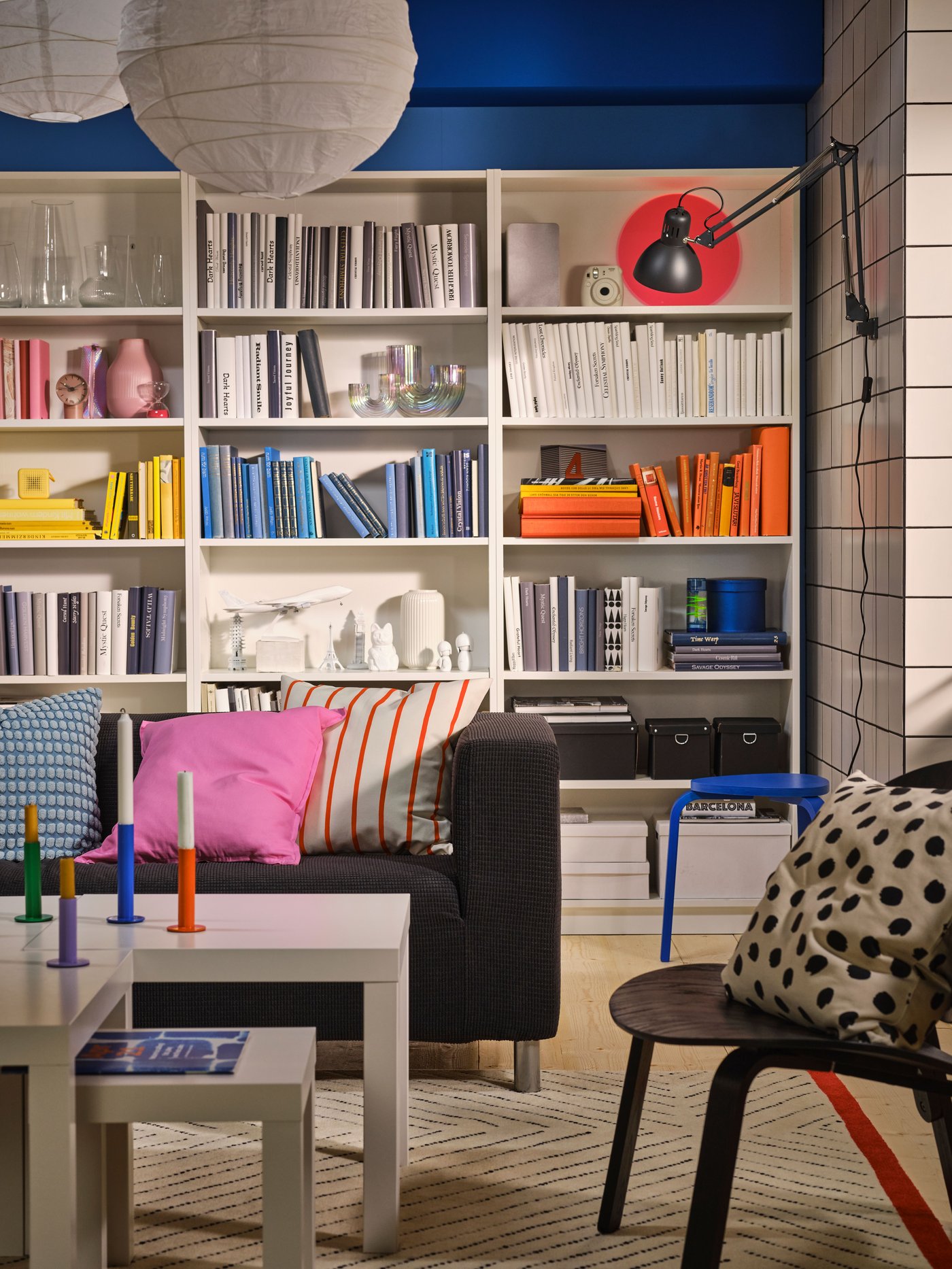 In front of a blue wall, there are three white shelves filled with books. In front of the shelves, there is a black sofa and a brown chair to the side.
