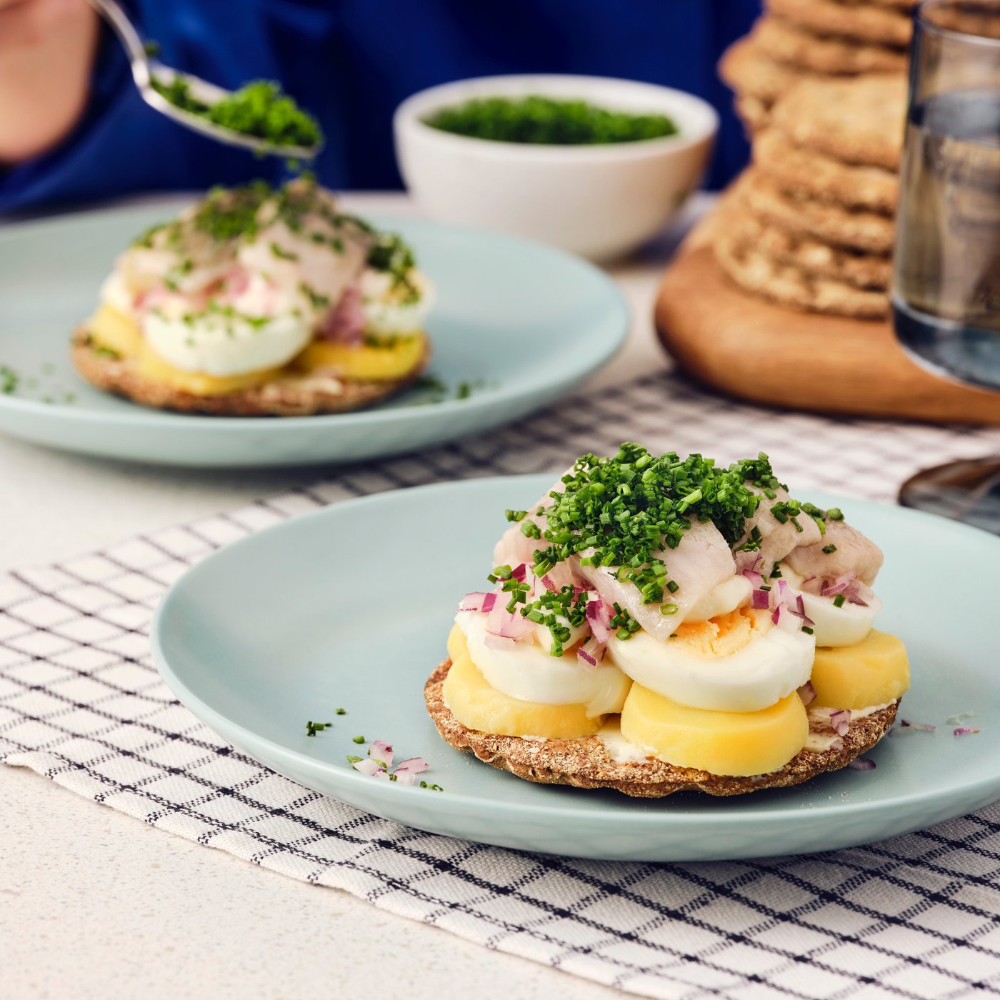 Two open sandwiches with KNÄCKEBRÖD RÅG rye crispbread, eggs, SILL DILL marinated herring, and chives on blue plates.