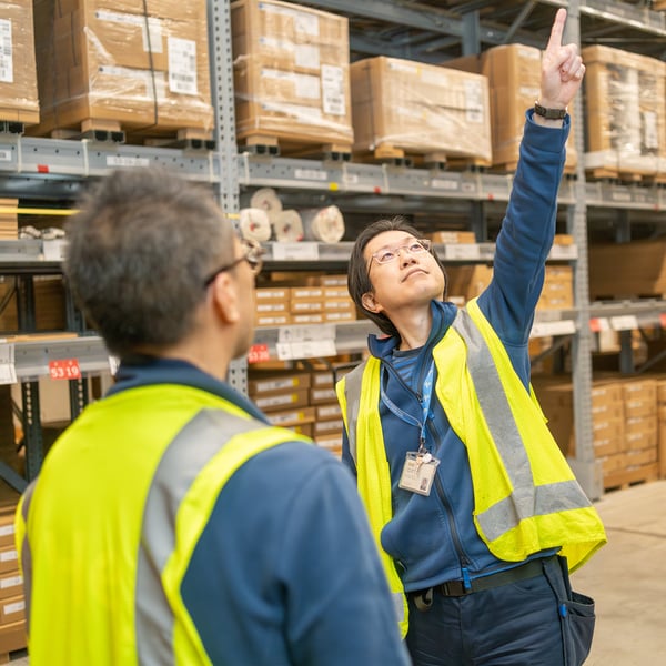 セルフサービスエリアで、反射ベストを着たイケアのコワーカー2人が高い棚の在庫を指さして確認している
