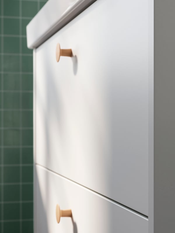 A bathroom with mint green tiles and a white HAVBÄCK wash-stand with two drawers and knobs in solid beech.