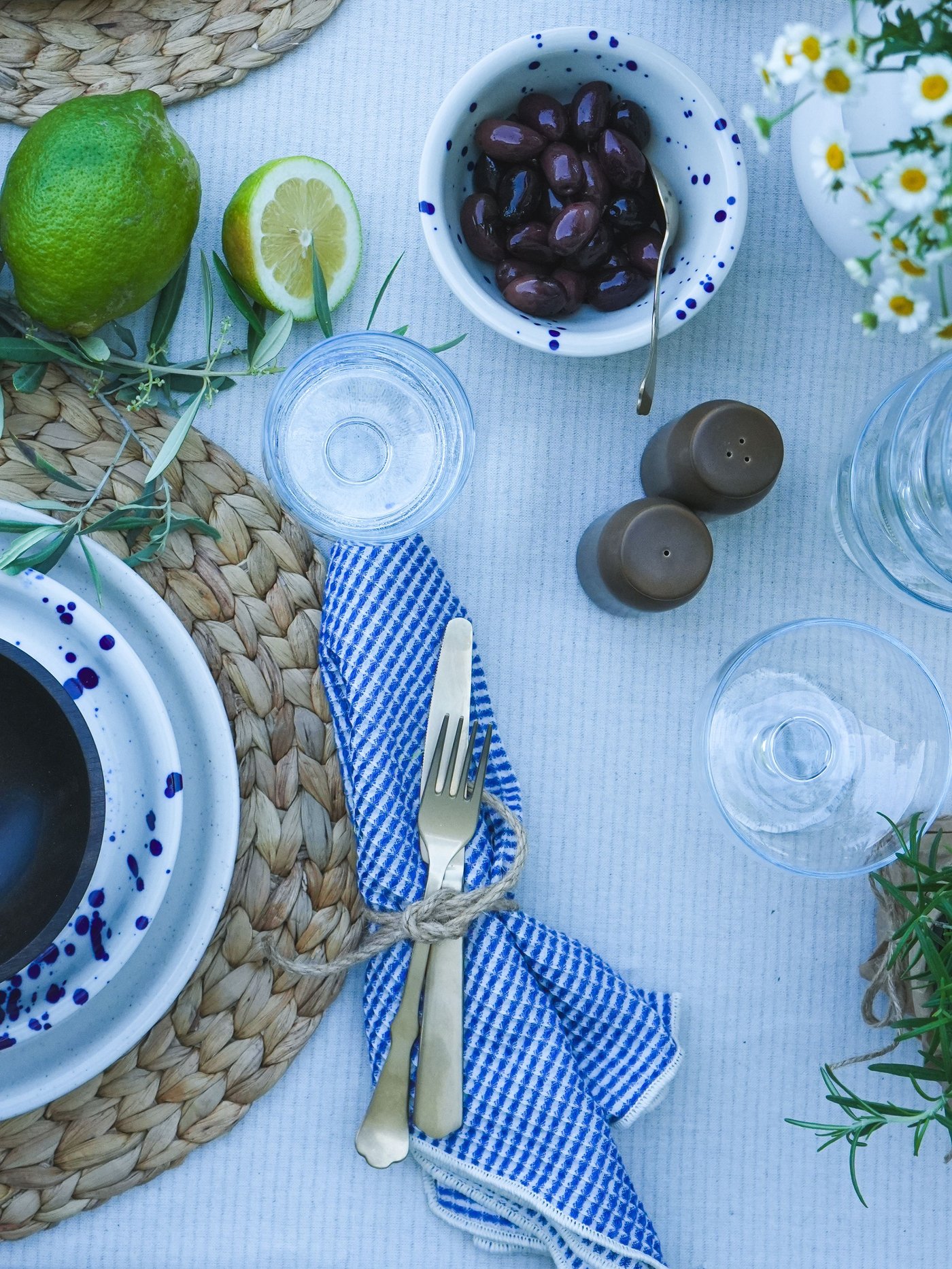 Une table dressée avec des couverts rustiques, un torchon de cuisine VÄLVÅRDAD bleu et blanc, un bol rempli d'olives, des citrons frais et des décorations florales.