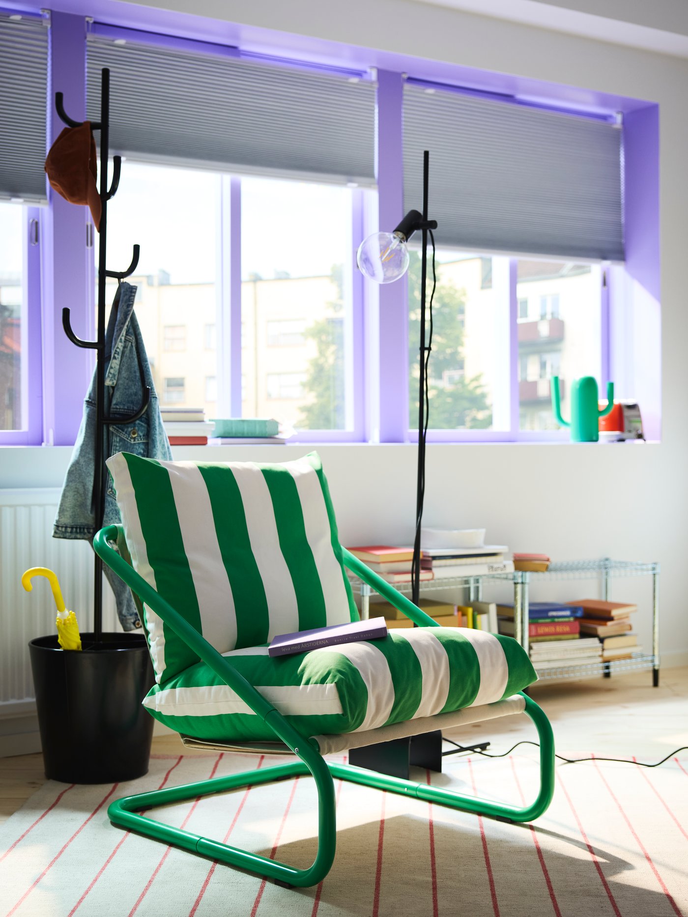 A green/white striped ÖNNESTAD armchair in the middle of a room with big windows and ÄLGANÄS hat and coat stand behind.