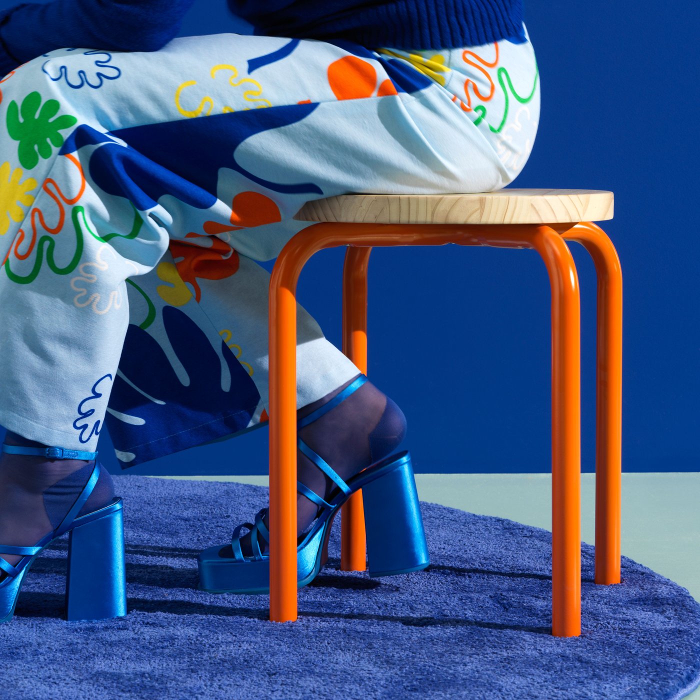 A person in trousers with a leaf pattern sits on a DOMSTEN stool with orange legs.