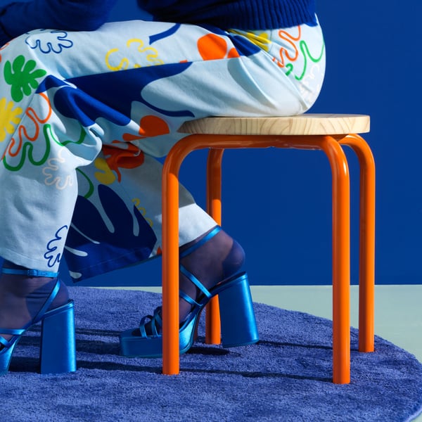 A person in trousers with a leaf pattern sits on a DOMSTEN stool with orange legs.