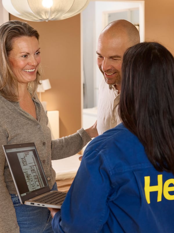 Standing in a showroom with light brown walls, an IKEA interior designer is showing two of her clients interior drawings on a laptop.