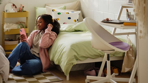 Une jeune fille assise par terre adossée à un lit avec textile de lit vert clair regarde son téléphone portable rose.
