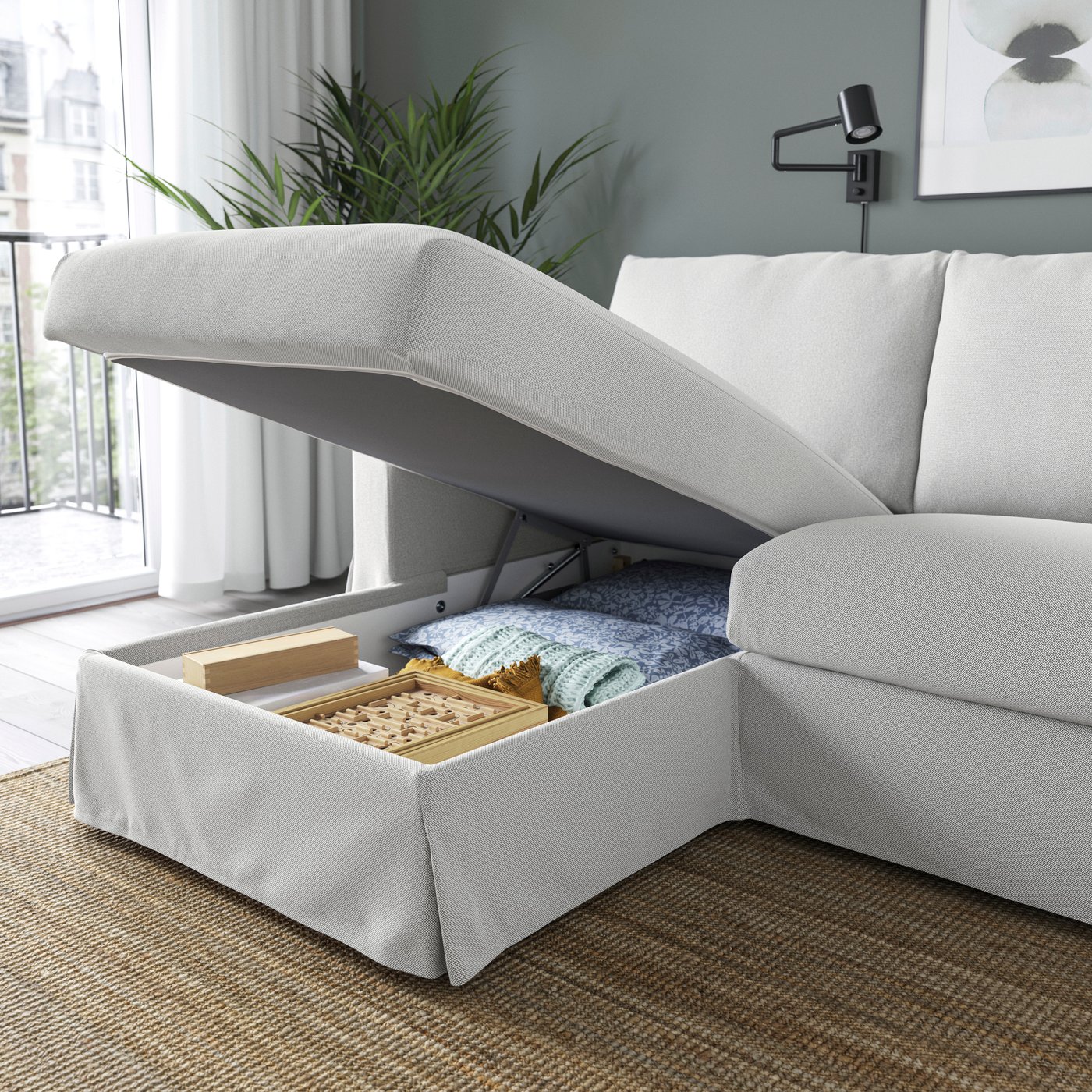 Divano HYLTARP 3 posti con chaise-longue a sinistra, rivestimento TALLMYRA bianco/nero, con vano contenitore sotto la seduta aperta che mostra coperte e accessori riposti.