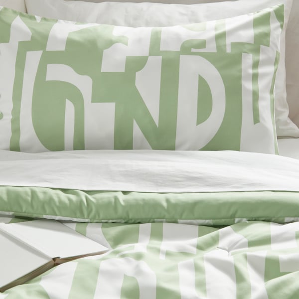 A light green and white comforter on a bed with a white notebook sitting face down on the bed.