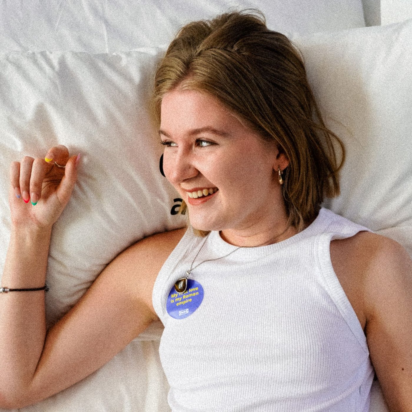 Izzy liegt auf einem Bett in einem weissen T-Shirt. Sie schaut nach rechts und lächelt, während sie ihre rechte Hand an ihr Gesicht hält.