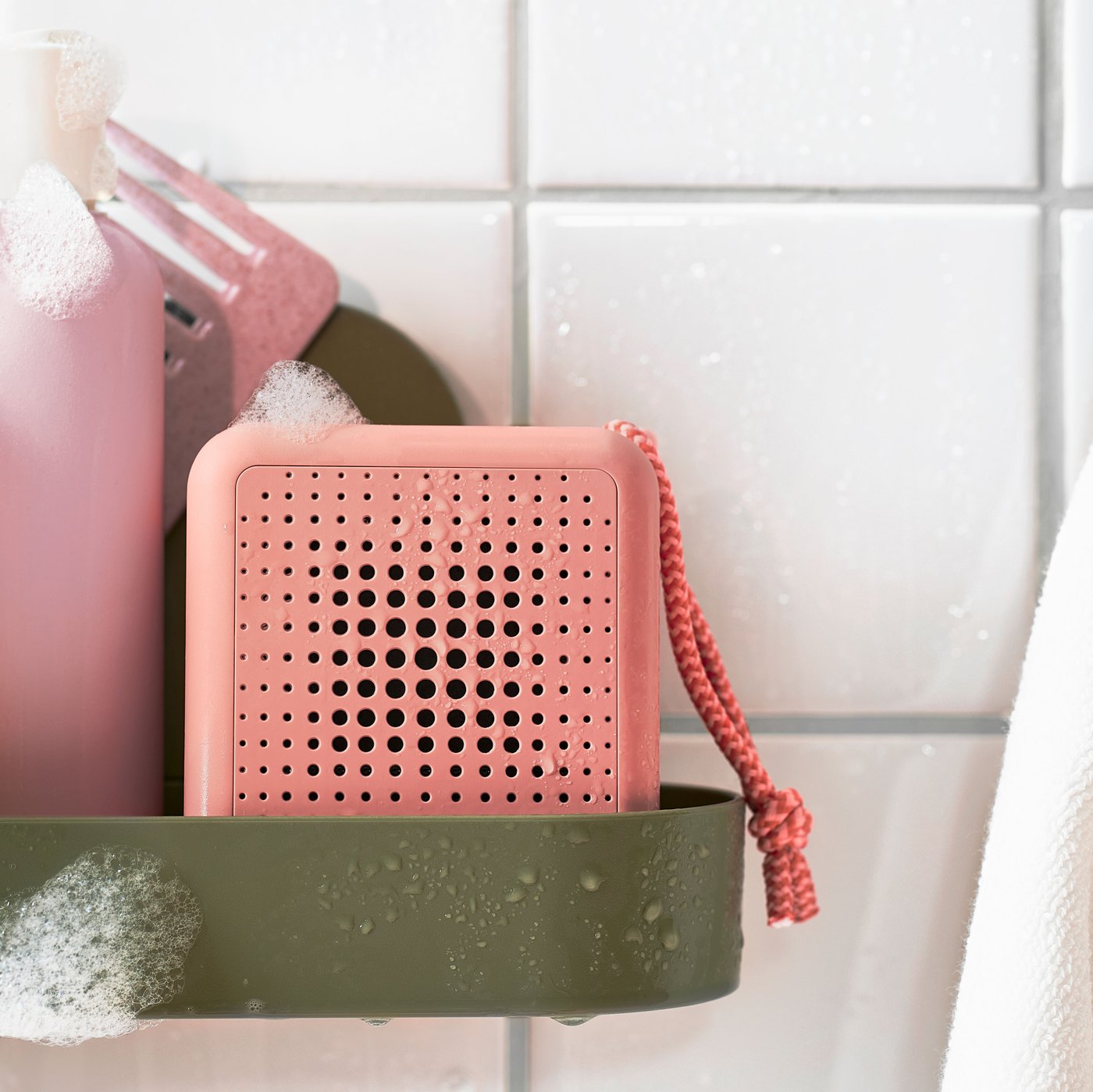 Una cassa Bluetooth portatile VAPPEBY impermeabile rosa è appoggiata su una mensola con ventosa ÖBONÄS grigio-verde in una doccia.