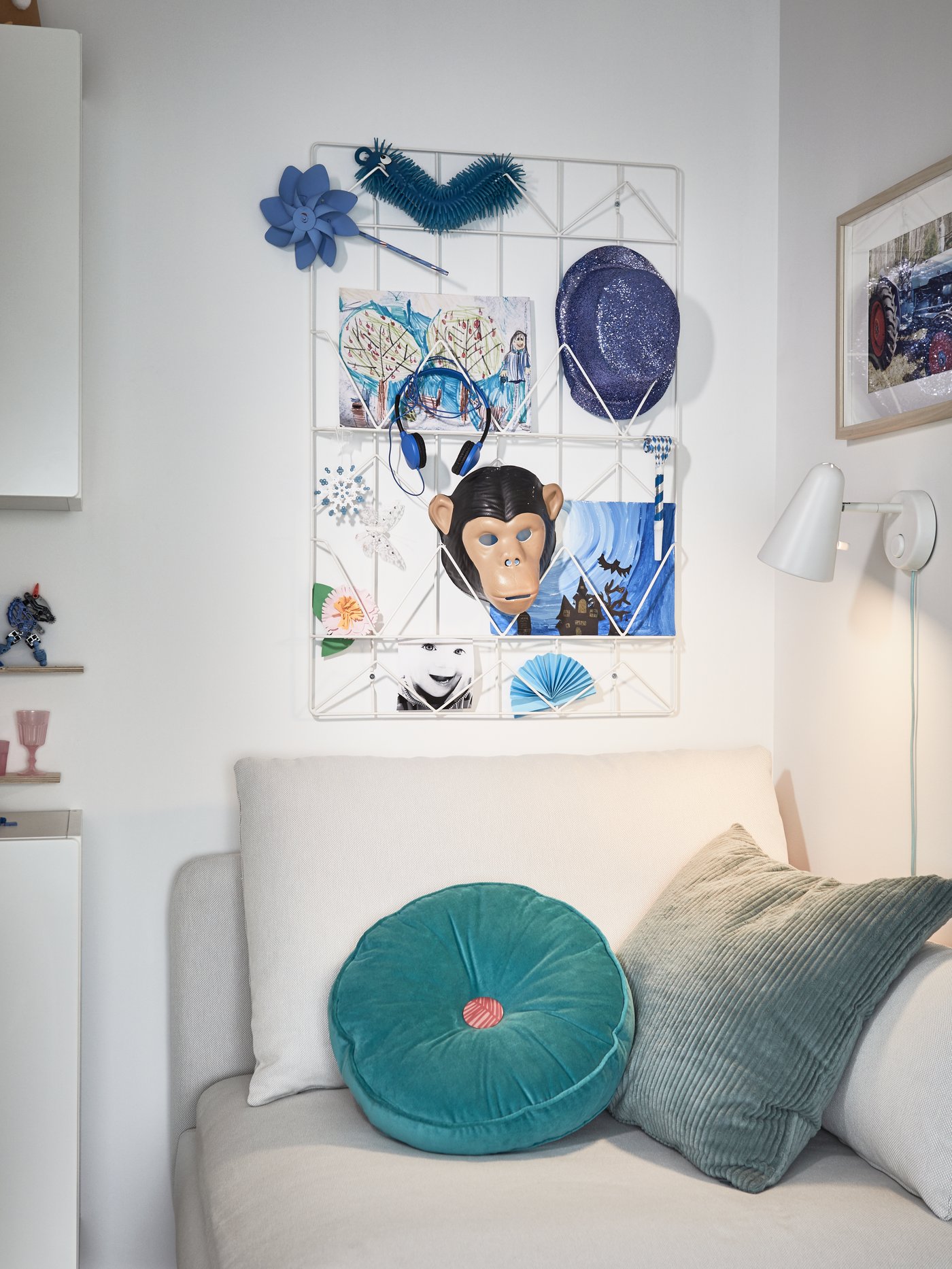 ホワイトのワイヤー製のメモボードに、ブルーの絵やアクセサリーがディスプレイされている。ベージュのソファ用モジュール、ターコイズのクッション、ホワイトのウォールランプ。