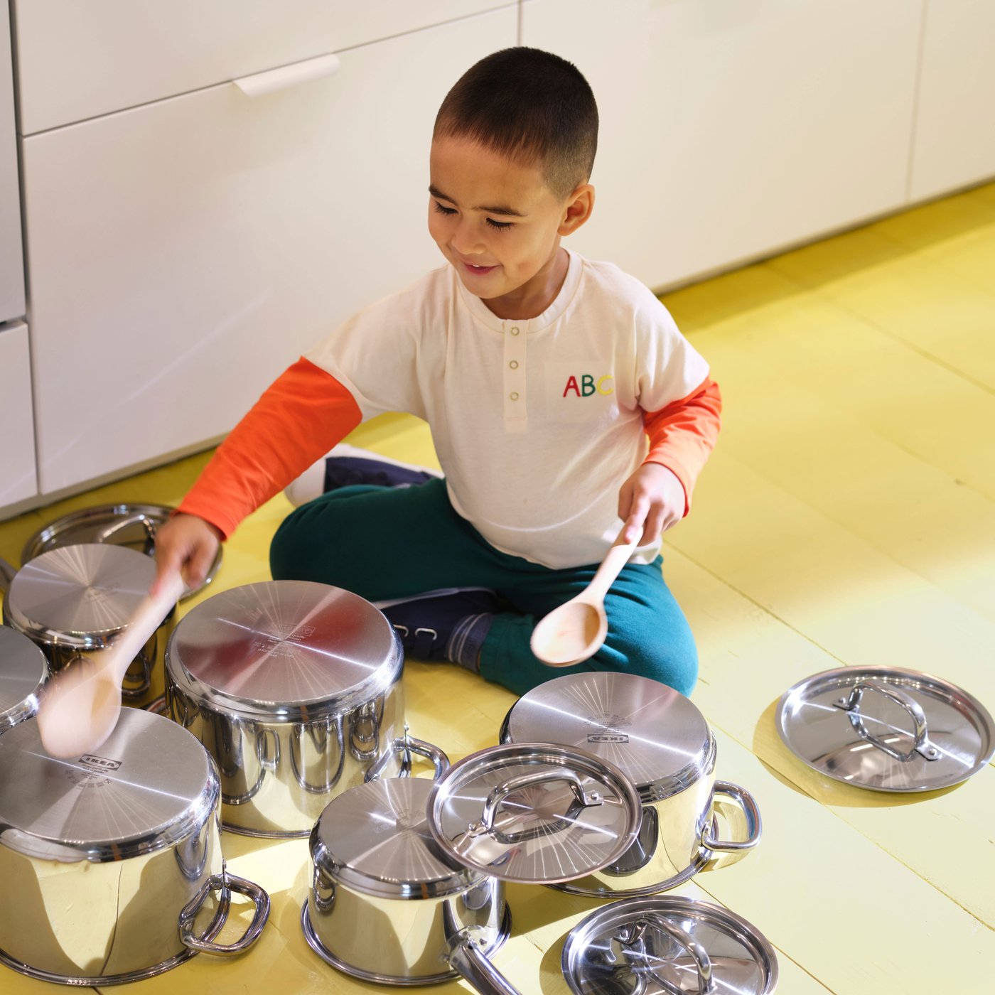 A little boy in a white T‑shirt with orange sleeves is sitting on the floor, playing with two ladles on upside‑down pots and lids.