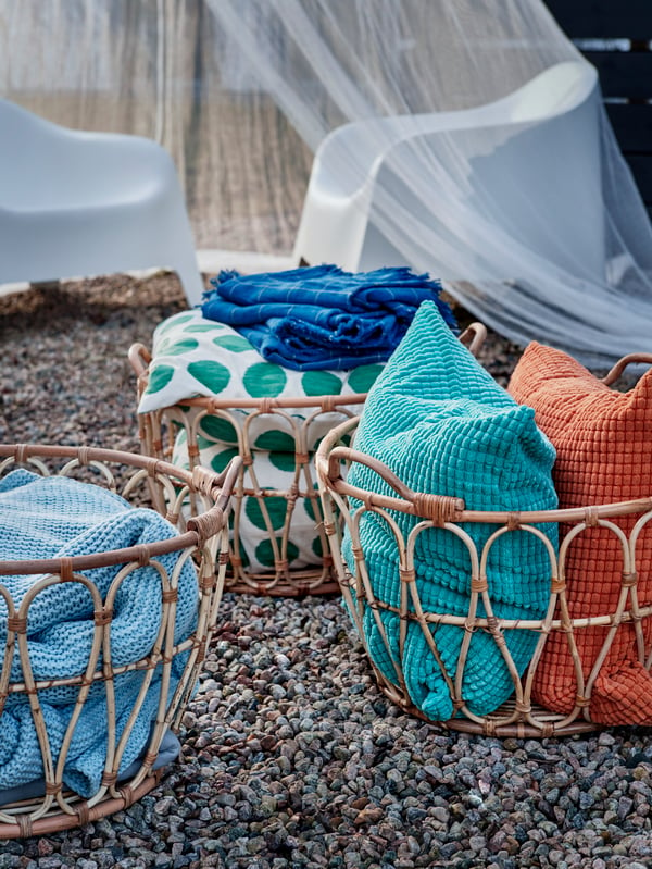 Three SNIDAD rattan baskets on a gravel surface, each containing neatly folded textiles.