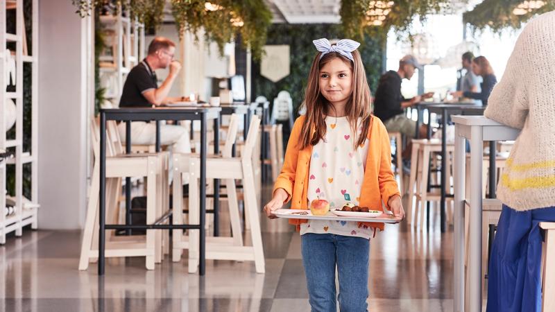 Ein Kind steht im IKEA Restaurant und hält ein Tablett mit Speisen. Dabei lächelt es.