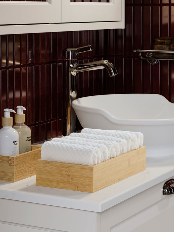 Un lavabo amb tovalloles de mà GULLVIAL blanques perfectament plegades en una safata de bambú al costat de pots de sabó líquid i crema de mans.
