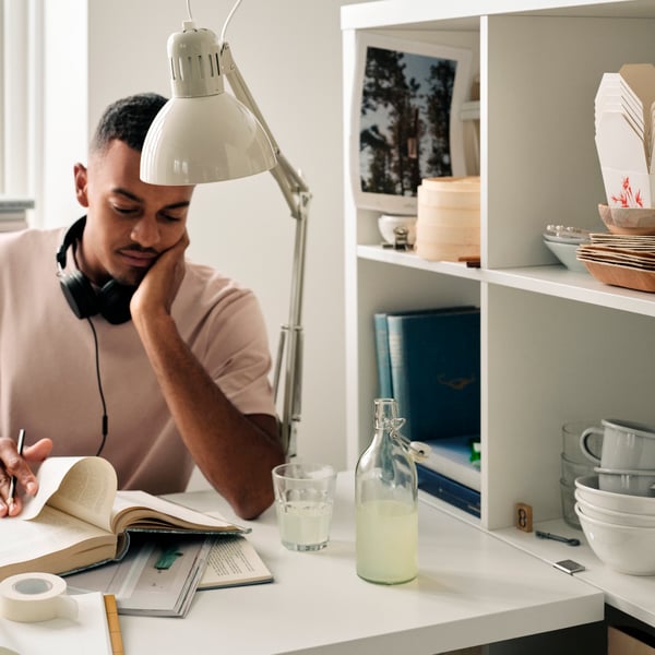 A man reading a book with headphones around his shoulders, sitting by a table with a white KALLAX shelving unit beside it.