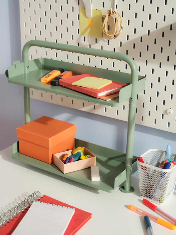 A light grey-green RELATERA desktop shelf on a white desk with stationery on it, and a white SKÅDIS pegboard behind it.